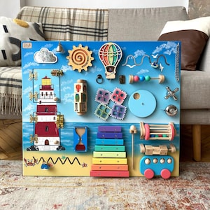 Può includere: Un pannello di attività in legno colorato con diverse texture e attività per bambini. Presenta un faro, un sole, una mongolfiera, uno xilofono, un'auto, un set di ingranaggi, una porta con un chiavistello, una nuvola, una campana, un granchio, una stella marina e una conchiglia.