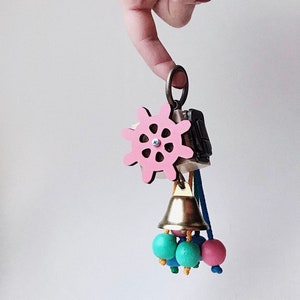 May include: A pink wooden toy with a steering wheel, a bell, and colorful beads. The toy is hanging from a metal ring.