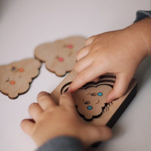 May include: A child's hand places a wooden cloud puzzle piece into a matching cutout. The puzzle piece has a smiling face with blue eyes and eyelashes. The puzzle is made of natural wood and has a dark brown border.