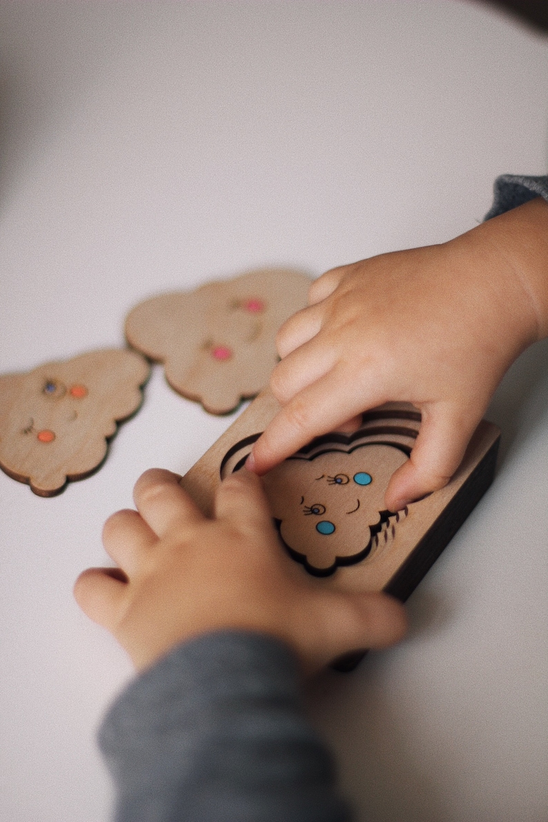 May include: A child's hand places a wooden cloud puzzle piece into a matching cutout. The puzzle piece has a smiling face with blue eyes and eyelashes. The puzzle is made of natural wood and has a dark brown border.