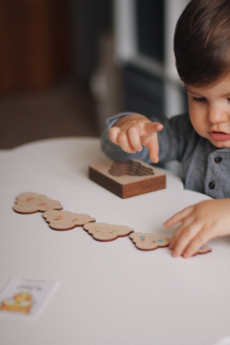 May include: A child plays with a wooden toy set featuring colorful cloud-shaped pieces. The pieces have different facial expressions and are arranged in a line on a white table.