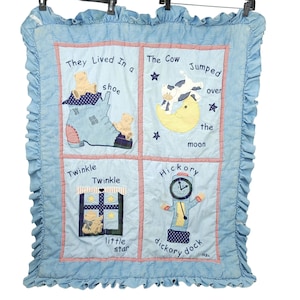 May include: A light blue baby quilt with four squares featuring nursery rhyme illustrations. The quilt includes a shoe with teddy bears, a cow jumping over the moon, a window with stars, and a clock. The edges are trimmed with a ruffled border.