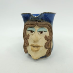 May include: A ceramic mug shaped like a face with brown hair, blue eyes, and a blue hat. The mug is hand-painted and has a whimsical design.