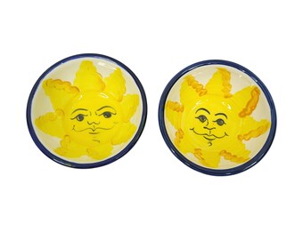 May include: Two small ceramic bowls with a yellow sun face design. The sun has a blue and black outline and a yellow face with black eyes and a smile.