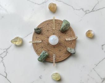 May include: A circular wooden board with a geometric pattern, surrounded by various crystals and stones. The arrangement includes a central white sphere, several clear quartz crystals, and green and yellow stones, creating a symmetrical display.