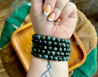 May include: A stack of dark green beaded bracelets. The bracelets are made of round beads with a speckled pattern. The bracelets are worn on a wrist with a tattoo. The nails are painted with a white and gold design.