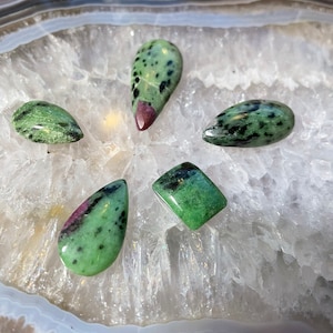 May include: Five green and black gemstone cabochons with a pink hue, displayed on a white agate geode slice. The stones are various shapes, including teardrop, oval, and square.