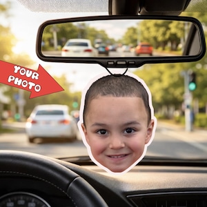 May include: A car air freshener featuring a smiling child's face, hanging from a rearview mirror. The text "YOUR PHOTO" is displayed on a red arrow pointing towards the air freshener. The background shows a street scene with cars.