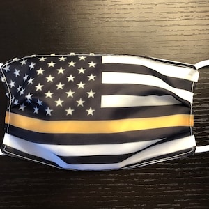 Puede incluir: Una mascarilla facial negra y blanca con una fina franja amarilla en el medio. La mascarilla presenta un diseño de la bandera estadounidense con una fina franja amarilla en el medio.