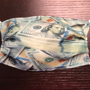 May include: A white face mask with a pattern of US dollar bills. The mask has white elastic ear loops.