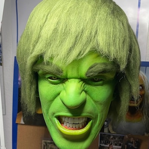 Peut inclure: Un masque vert sculpté représentant un personnage à l'expression en colère. Le masque présente une peau verte, des sourcils froncés et une bouche grande ouverte révélant des dents blanches et un intérieur jaune-vert. Les cheveux sont vert clair, coiffés en coupe hirsute.