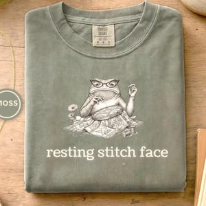 May include: A sage green t-shirt featuring a graphic of a frog wearing glasses and sewing a quilt with a needle and thread. The text "resting stitch face" is printed in white below the illustration. The shirt has a Comfort Colors brand label.