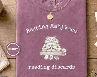 May include: A berry-colored crewneck t-shirt featuring a graphic of a frog wearing glasses behind a mahjong rack with the text Resting Mahj Face and reading discards. A gold-toned necklace with a round pendant rests on the neckline of the shirt.
