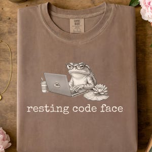 May include: A brown t-shirt featuring a graphic of a toad wearing glasses, typing on a laptop next to a coffee cup and a water lily. The text "resting code face" is printed in white below the illustration. The shirt has a crew neck and a label.