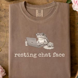 May include: A brown crewneck t-shirt featuring a graphic of a toad wearing glasses, typing on a laptop next to a coffee cup and a water lily. The text "resting chat face" is printed in white below the illustration, with a brand label at the neck.