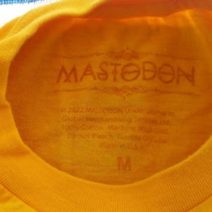 Pode incluir: Camiseta amarela com um logotipo preto MASTODON estampado na frente. A etiqueta no interior da camiseta diz "2022 MASTODON Under license Global Merchandising Services, Ltd. 100% Cotton Machine Wash Cold" e tem tamanho M.