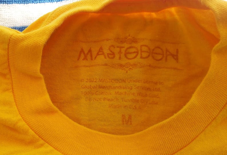 Pode incluir: Camiseta amarela com um logotipo preto MASTODON estampado na frente. A etiqueta no interior da camiseta diz "2022 MASTODON Under license Global Merchandising Services, Ltd. 100% Cotton Machine Wash Cold" e tem tamanho M.