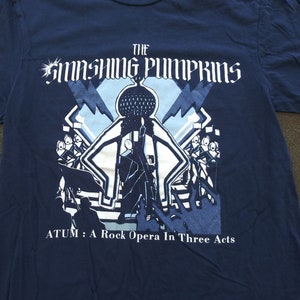 May include: A navy blue t-shirt with a graphic of a band, The Smashing Pumpkins, with the text "ATUM: A Rock Opera In Three Acts".