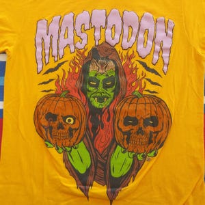 Pode incluir: Uma camiseta amarela com uma estampa gr&aacute;fica de uma mulher de pele verde com presas segurando duas ab&oacute;boras de Halloween. O texto "MASTODON" est&aacute; impresso em roxo acima da mulher.