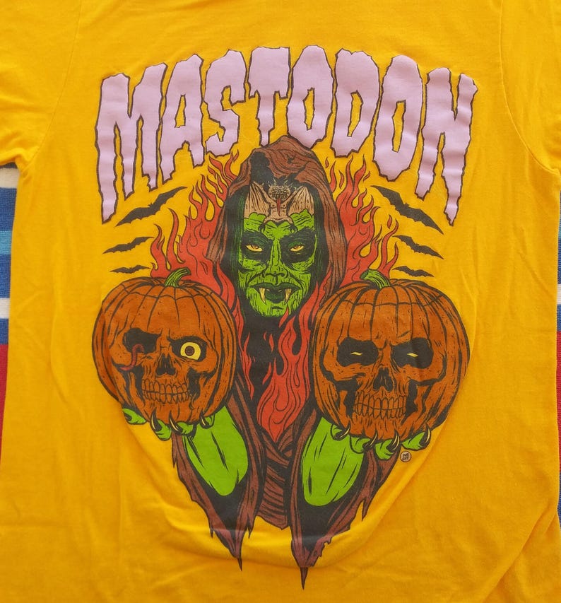 Pode incluir: Uma camiseta amarela com uma estampa gr&aacute;fica de uma mulher de pele verde com presas segurando duas ab&oacute;boras de Halloween. O texto "MASTODON" est&aacute; impresso em roxo acima da mulher.
