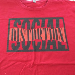 May include: A red t-shirt with a black and orange graphic that reads "SOCIAL DISTORTION". The text is in a bold, distressed font, with the word "DISTORTION" in a contrasting orange color. The shirt is made of a soft material.