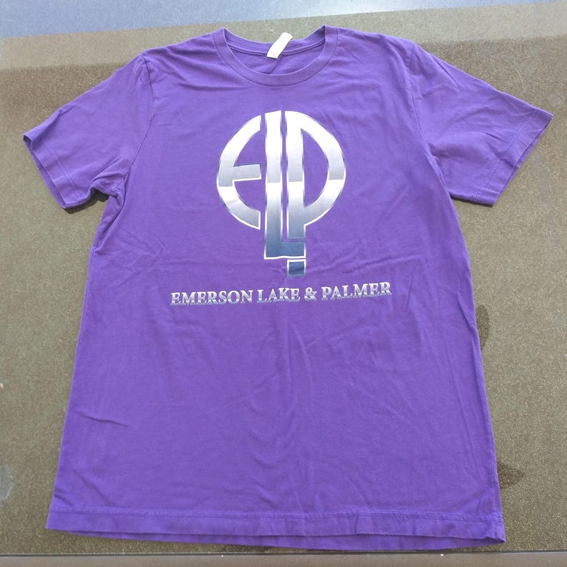 May include: Purple t-shirt with a silver circle logo featuring the letters "ELP" and the text "Emerson Lake & Palmer" below the logo.