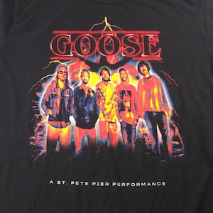 May include: Black t-shirt with the band name "Goose" in a red, stylized font above a graphic of the band members. The text "A St. Pete Pier Performance" is printed below. The band members are wearing jackets and shirts.