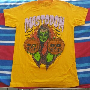 Pode incluir: Camiseta amarela com um gr&aacute;fico de um monstro verde segurando duas ab&oacute;boras de Halloween. O texto "MASTODON" est&aacute; impresso em roxo acima do monstro.