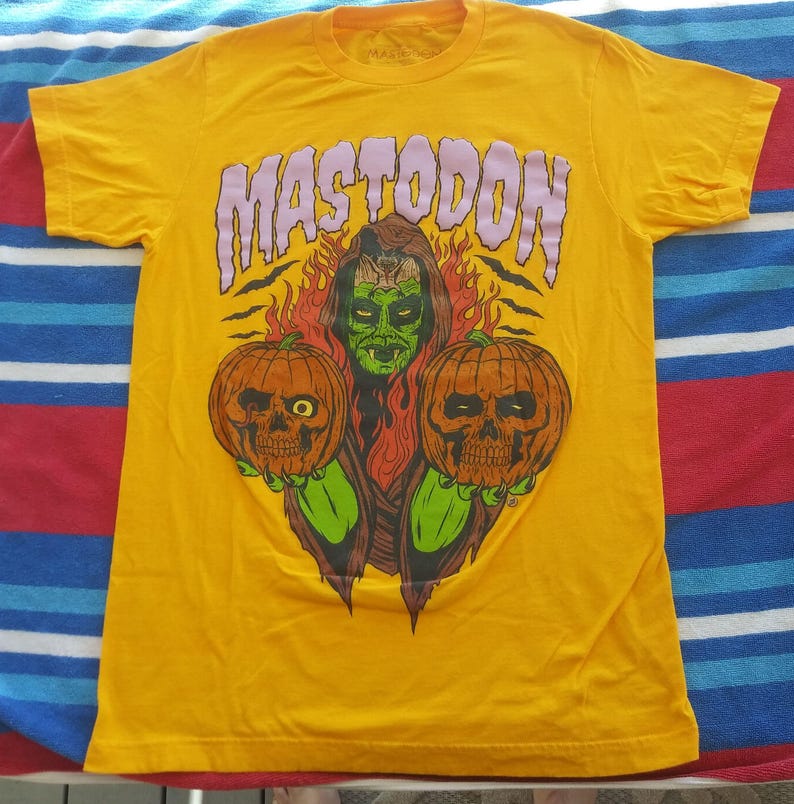 Pode incluir: Camiseta amarela com um gr&aacute;fico de um monstro verde segurando duas ab&oacute;boras de Halloween. O texto "MASTODON" est&aacute; impresso em roxo acima do monstro.