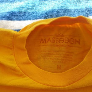 Pode incluir: Camiseta amarela com um logotipo preto "Mastodon" estampado na frente. A camiseta tem gola redonda e mangas curtas. A etiqueta no interior da camiseta diz "Mastodon" e inclui informa&ccedil;&otilde;es sobre o tamanho.