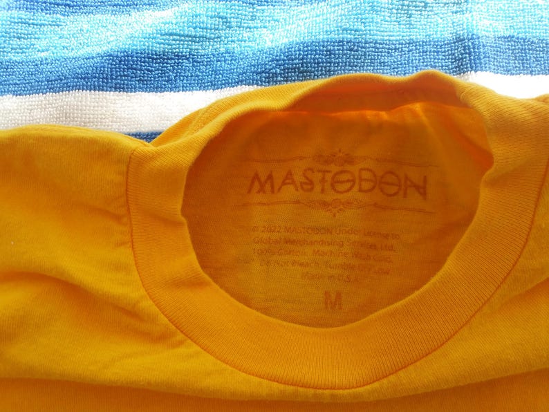 Pode incluir: Camiseta amarela com um logotipo preto "Mastodon" estampado na frente. A camiseta tem gola redonda e mangas curtas. A etiqueta no interior da camiseta diz "Mastodon" e inclui informa&ccedil;&otilde;es sobre o tamanho.