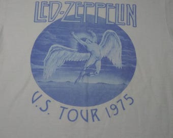 May include: White t-shirt featuring a blue graphic of a winged figure within a blue circle. The text "LED ZEPPELIN" is above the graphic, and "U.S. TOUR 1975" is below. The shirt is made of a soft material.