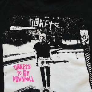 May include: Black t-shirt featuring a graphic of a person holding a guitar. The image includes the text "TICKETS" at the top and "TICKETS TO MY DOWNFALL" in pink below the person.