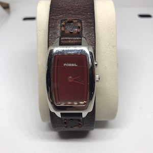 Puede incluir: Un reloj Fossil con una esfera rectangular de color granate y una caja plateada. El reloj tiene una correa de cuero marrón. La palabra "Fossil" está impresa en la esfera. El reloj se muestra sobre un soporte blanco.