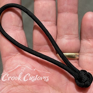 以下が含まれることがあります： 黒いパラコードのキーホルダーで、端に結び目があります。キーホルダーは手に持たれています。