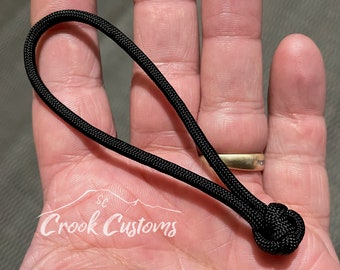 May include: Black paracord keychain with a knot at the end. The keychain is held in a hand.