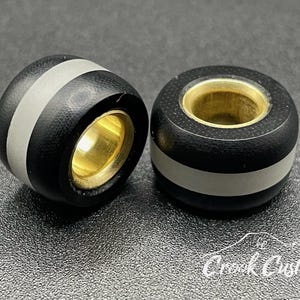K&ouml;nnte beinhalten: Zwei schwarze und silberne Perlen mit goldfarbenem Innenraum. Die Perlen haben eine strukturierte Oberfl&auml;che und einen wei&szlig;en Streifen um die Mitte. Der Text "Crook Customs" ist unten rechts sichtbar.