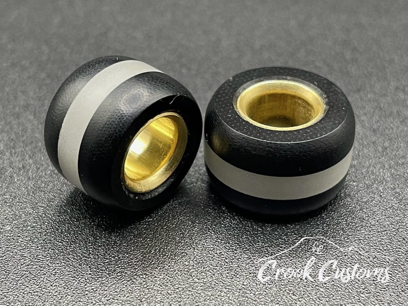 K&ouml;nnte beinhalten: Zwei schwarze und silberne Perlen mit goldfarbenem Innenraum. Die Perlen haben eine strukturierte Oberfl&auml;che und einen wei&szlig;en Streifen um die Mitte. Der Text "Crook Customs" ist unten rechts sichtbar.