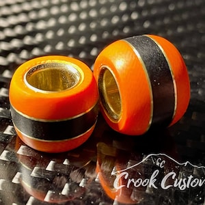 May include: Two orange and black wooden beads with a gold accent. The beads have a smooth, polished finish and are designed for use in jewelry making.