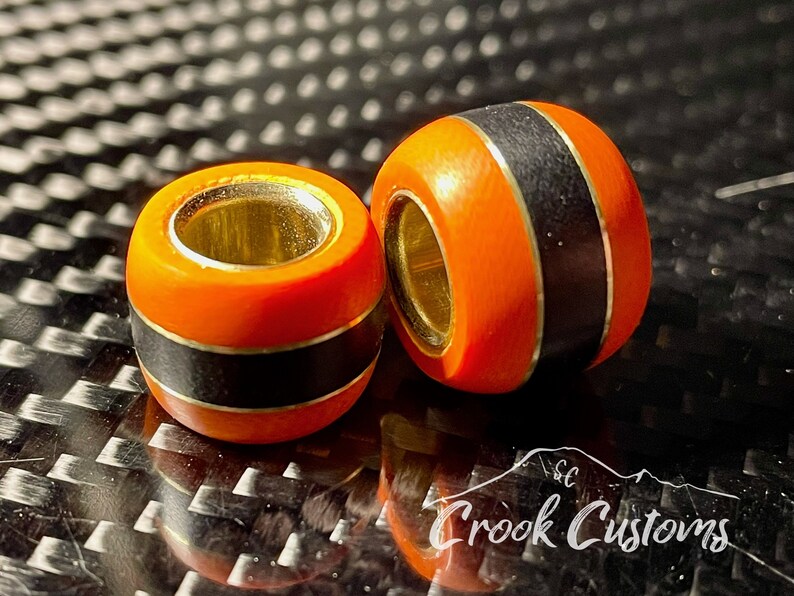 May include: Two orange and black wooden beads with a gold accent. The beads have a smooth, polished finish and are designed for use in jewelry making.