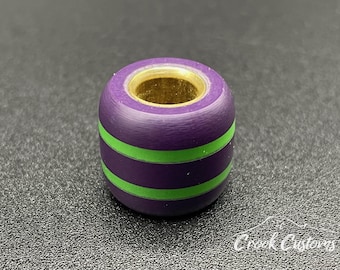 May include: A cylindrical bead featuring a purple exterior with two horizontal lime green stripes and a metallic gold-toned inner core. The bead has a smooth, matte finish and a hollow center, designed for threading onto a cord or chain for jewelry.