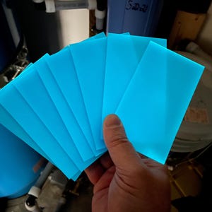 May include: A hand holds a fan of bright blue, translucent, rectangular sheets. The sheets are evenly spaced, fanning out to reveal a glowing effect. The background includes plumbing and storage containers.