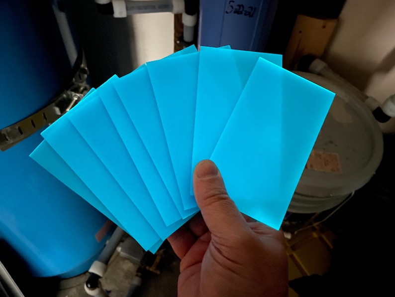 May include: A hand holds a fan of bright blue, translucent, rectangular sheets. The sheets are evenly spaced, fanning out to reveal a glowing effect. The background includes plumbing and storage containers.