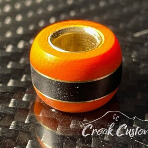 May include: Orange and black bead with a gold metal ring. The bead has a black band with a thin gold line on either side.