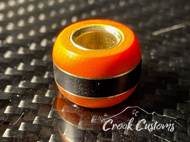 May include: Orange and black bead with a gold metal ring. The bead has a black band with a thin gold line on either side.
