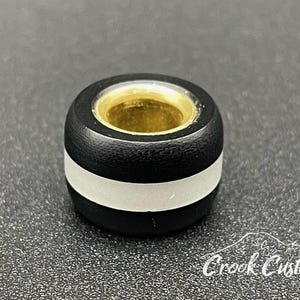 K&ouml;nnte beinhalten: Eine schwarz-silberne Perle mit goldfarbenem Innenraum. Die Perle hat ein silbernes Band in der Mitte und ist zylindrisch geformt. Die Perle liegt auf einer strukturierten Oberfl&auml;che.