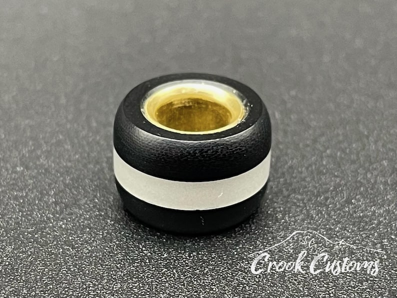 K&ouml;nnte beinhalten: Eine schwarz-silberne Perle mit goldfarbenem Innenraum. Die Perle hat ein silbernes Band in der Mitte und ist zylindrisch geformt. Die Perle liegt auf einer strukturierten Oberfl&auml;che.