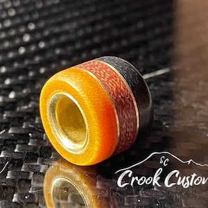 May include: A round, multi-colored bead with a gold ring around the center hole. The bead is made of resin and has a black, red, and orange color scheme. The bead is sitting on a black and white textured surface.