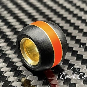 May include: A black and orange bead with a gold-colored interior. The bead has a textured surface and a bright orange stripe around the center. The bead is set against a dark, patterned background.