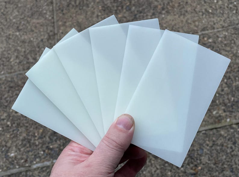 May include: Six translucent white sheets of paper are fanned out in a hand. The sheets are slightly textured and have a subtle sheen.